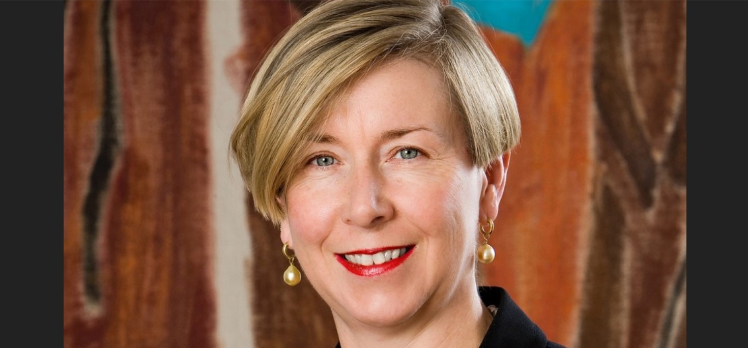 Portrait of a smiling middle-aged woman with short blonde hair, wearing red lipstick, black attire, and pearl earrings against a multi-colored abstract background.