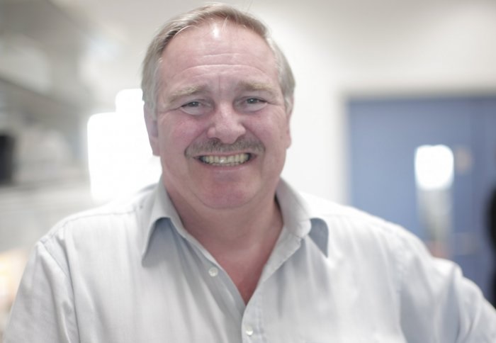 A man smiling broadly in a white shirt, standing in a brightly lit room.