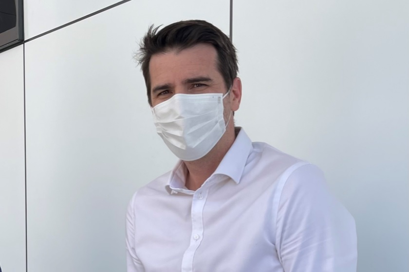 A man wearing a white shirt and a medical face mask standing in front of a light-colored background.