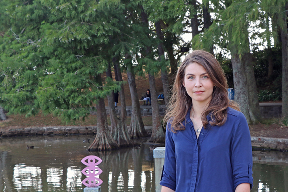 A woman in a blue shirt standing in front of a pond surrounded by dense trees.