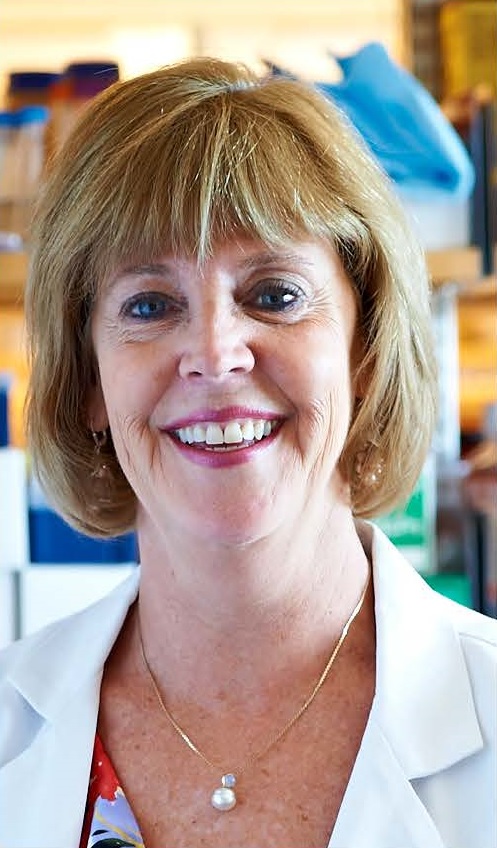 A portrait of a smiling woman with short blonde hair, wearing a white lab coat and a necklace.