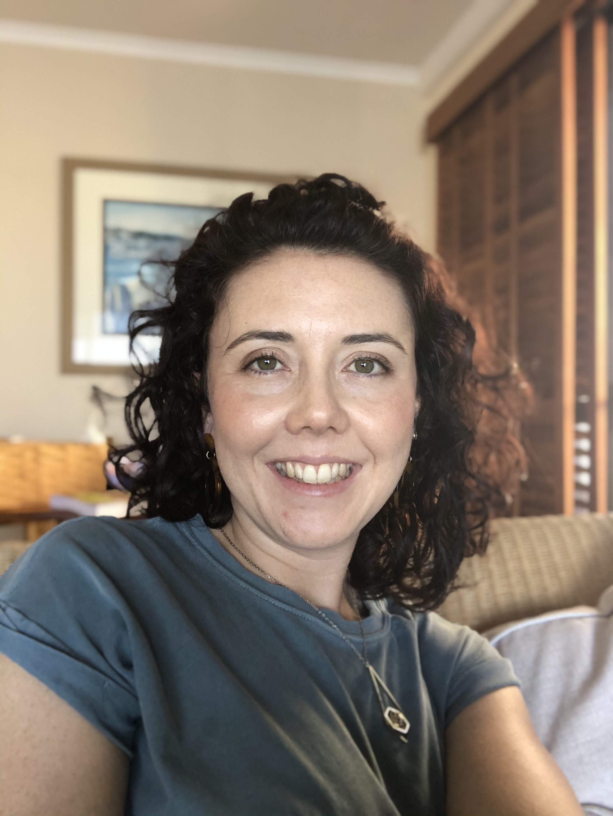 A smiling woman with curly dark hair wearing a blue t-shirt, seated indoors.
