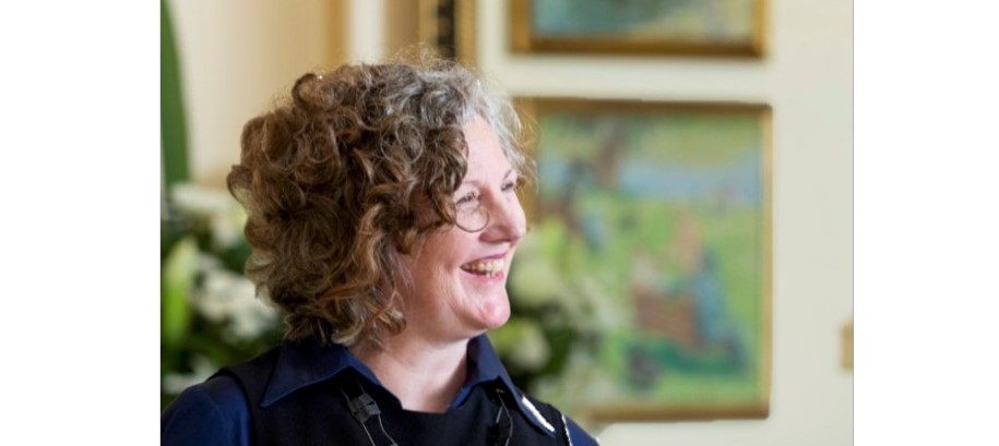 A woman with curly hair smiling and looking to the side in a room with a painting in the background.