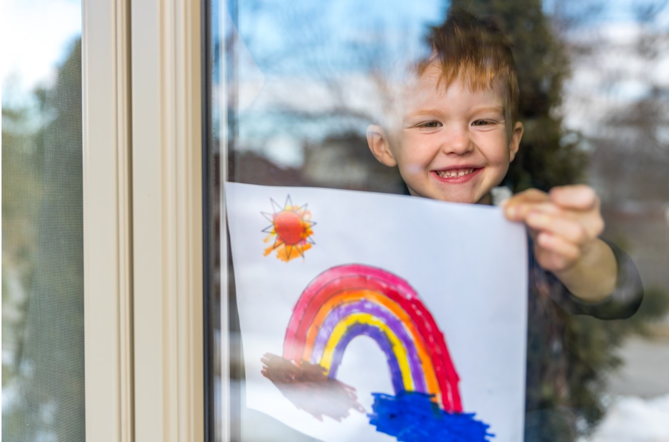 A young boy smiles as he shows off his colorful drawing of a rainbow and sun through a window.