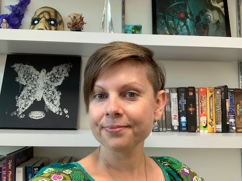 A woman with short hair smiling in front of a bookshelf filled with DVDs and various decorative items.