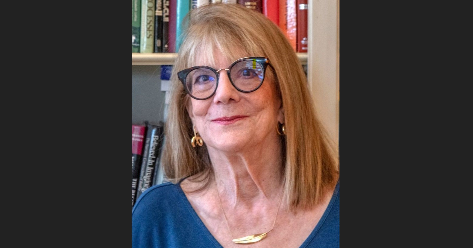 A senior woman with blonde hair and glasses, smiling in front of a bookshelf.
