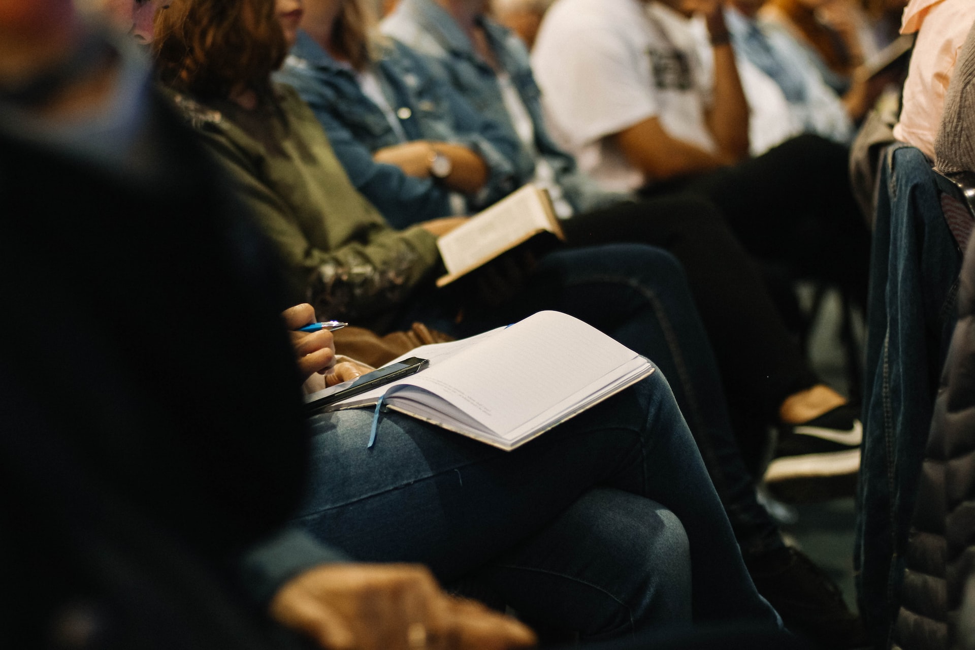 People seated in a lecture hall with notebooks on their laps, focused on writing notes.