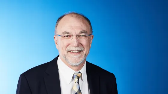 A smiling elderly man wearing glasses, a black suit, and a plaid blue and yellow tie, against a solid blue background.