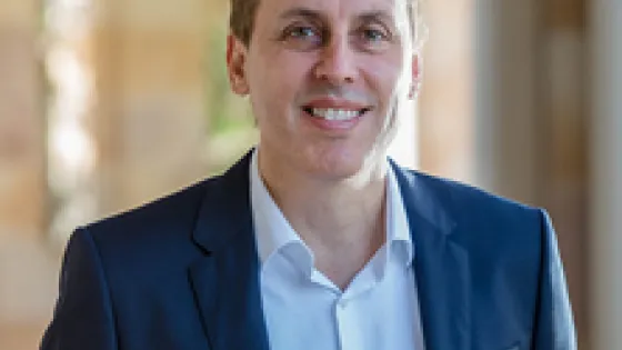 A smiling man in a dark suit jacket standing outdoors, with a softly blurred background.