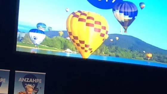A person standing at a podium in a conference hall, with a large screen behind them displaying an image of colorful hot air balloons and the text "ANZAHPE 2019."