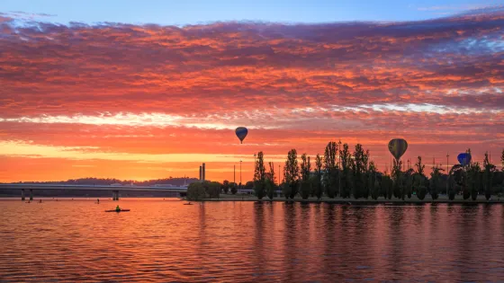 A vibrant sunset with cloud-streaked skies in shades of orange and blue over a calm lake, with hot air balloons floating in the distance and a kayaker on the water.