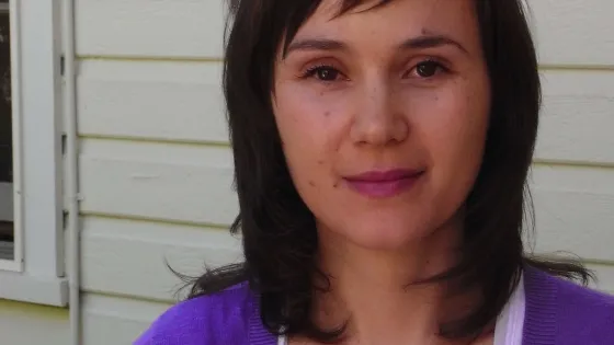 A woman with shoulder-length brown hair and a purple cardigan stands against a yellow house background.