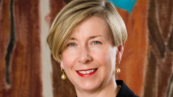 Portrait of a smiling middle-aged woman with short blonde hair, wearing red lipstick, black attire, and pearl earrings against a multi-colored abstract background.