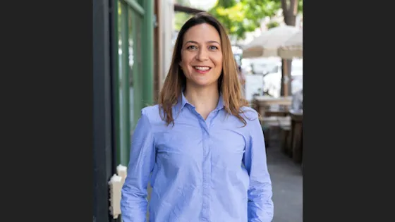 A woman in a blue shirt smiling while standing on a sidewalk next to a cafe.