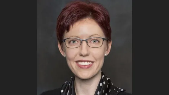 A woman with red hair and glasses, smiling, wearing a black and white polka dot scarf.