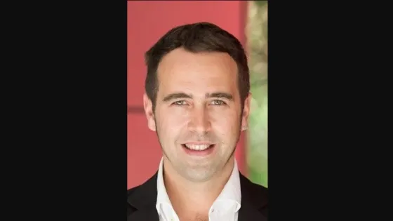 A portrait of a smiling man with dark hair, wearing a black suit, against a red background.