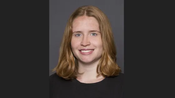 Portrait of a smiling young woman with shoulder-length red hair and a black top against a gray background.
