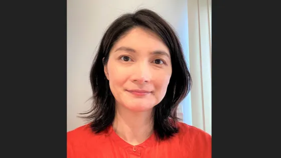 A middle-aged Asian woman with shoulder-length black hair, wearing a red blouse, smiling gently at the camera in an indoor setting.