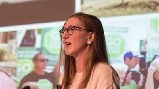 A woman with glasses speaking at a podium with a promotional banner in the background.