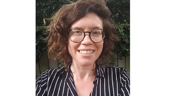 A woman with curly hair and glasses smiling in front of a greenery background, wearing a striped shirt.