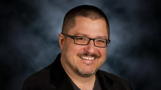 A smiling man with glasses and a beard, wearing a black jacket, against a blurred blue background.