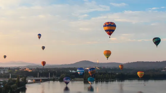 Colorful hot air balloons float over a serene lake and city during dawn.