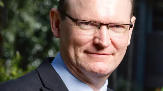 A portrait of a smiling man with neatly combed hair, wearing glasses, a dark suit, and a striped tie.
