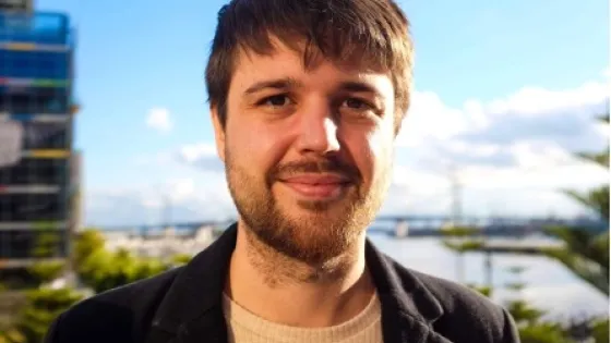 A man with short brown hair and a beard smiling at the camera, with a cityscape and clear sky in the background.