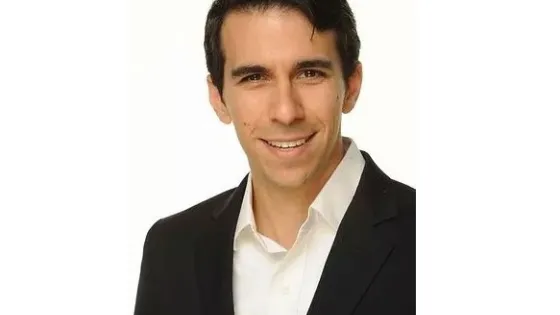 A man with dark hair, smiling, wearing a black blazer over a white shirt against a light background.