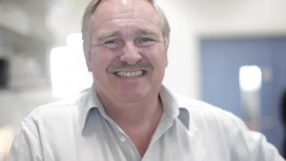 A man smiling broadly in a white shirt, standing in a brightly lit room.