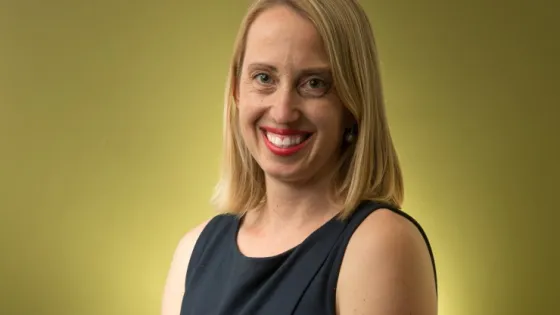 A smiling woman with blonde hair, wearing a sleeveless navy dress, against a greenish-yellow background.
