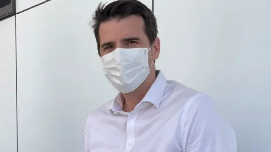 A man wearing a white shirt and a medical face mask standing in front of a light-colored background.