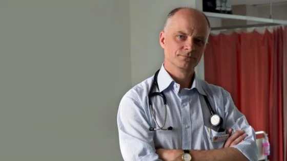 A bald man with a serious expression wearing a striped shirt, tie, and stethoscope stands with his arms crossed in a hospital setting.