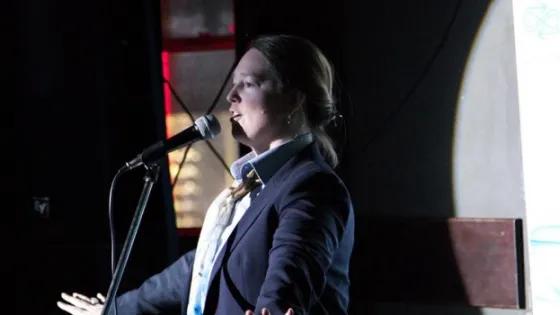 A woman speaks into a microphone on stage at a dimly lit event with a projection screen visible in the background.