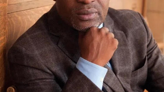 A man in a brown blazer and glasses sitting thoughtfully with his chin resting on his hand.