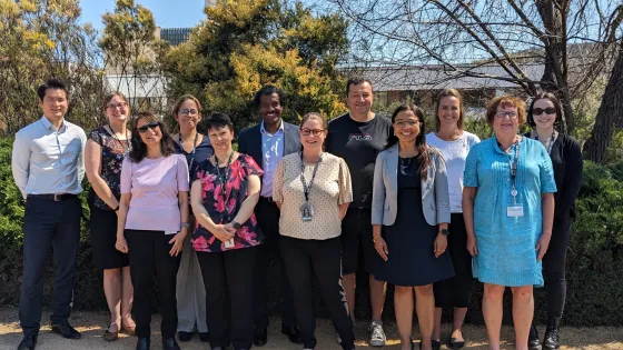 A group of eleven diverse professionals smiling for a photo outdoors on a sunny day.