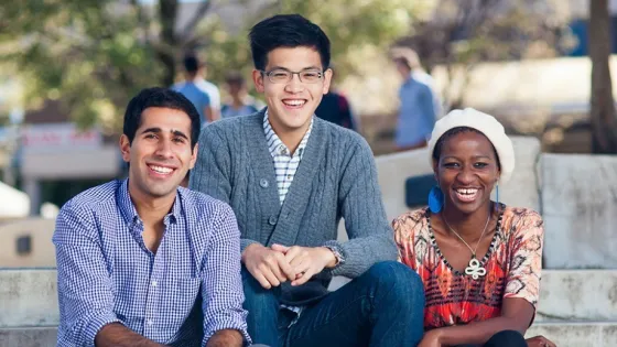 Three young adults, smiling and sitting together outdoors on a sunny day.