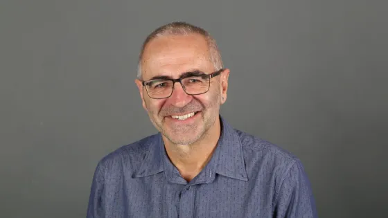 A smiling elderly man with glasses wearing a blue shirt against a gray background.
