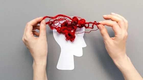 Hands arranging tangled red thread over a white paper cutout of a human head on a gray background.