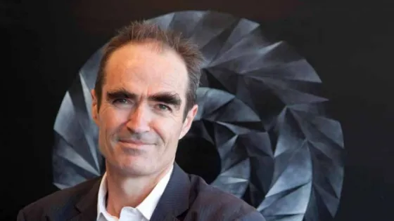 A man with dark hair and a slight smile wearing a dark suit stands in front of a geometric black and white artwork.