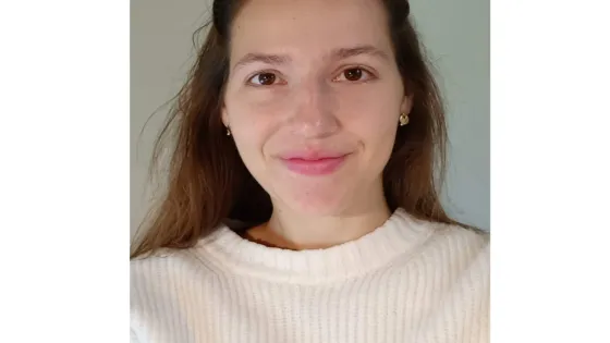 A young woman with brown hair wearing a white sweater smiles gently at the camera in a well-lit room.