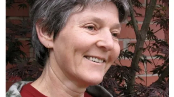 An older woman with short gray hair smiling gently, wearing a red sweater, with a backdrop of trees and a brick wall.