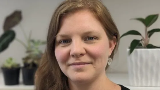 A woman with long brown hair smiling gently, wearing a green top, and standing in front of a bookshelf with plants.