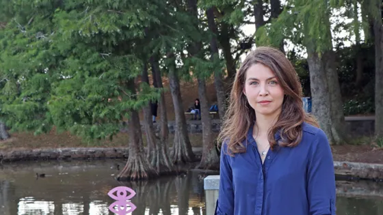 A woman in a blue shirt standing in front of a pond surrounded by dense trees.