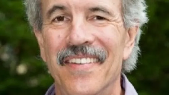 A portrait of an elderly man with a warm smile, sporting a neat gray mustache and wearing a purple shirt, with a blurred green background.