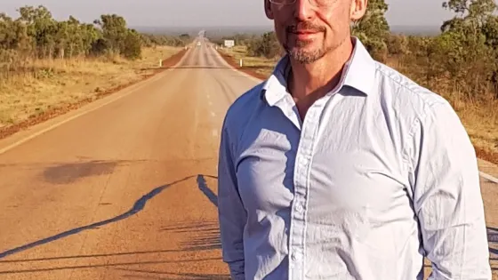 A man stands in the middle of a long, straight road stretching into the distance, wearing glasses and a semi-formal shirt.