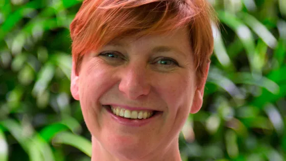 A woman with short red hair smiling against a backdrop of green foliage.