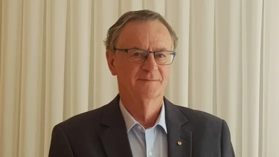 An older man wearing glasses and a dark suit stands in front of a curtain, smiling slightly at the camera.