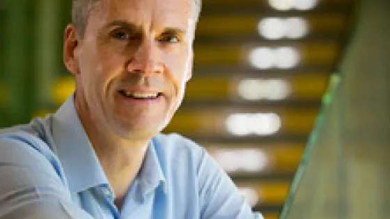 A man with grey hair, wearing a light blue shirt, smiling at the camera in a sunlit, green-toned architectural environment.