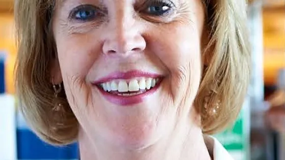A portrait of a smiling woman with short blonde hair, wearing a white lab coat and a necklace.